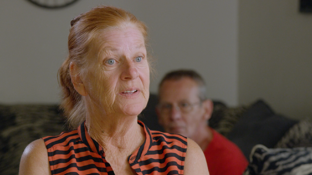 Image of an older woman speaking while her husband sits in the background.