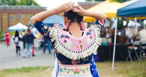 Image of a young Native girl wearing traditional tribal dress, seen from behind.