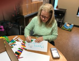 Image of an older woman sitting at a table writing a card that says "Merry Christmas!"