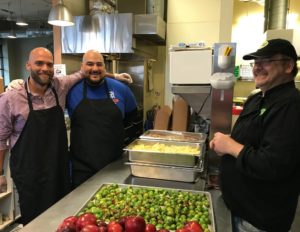 Image of three smiling men preparing meals in an industrial kitchen.