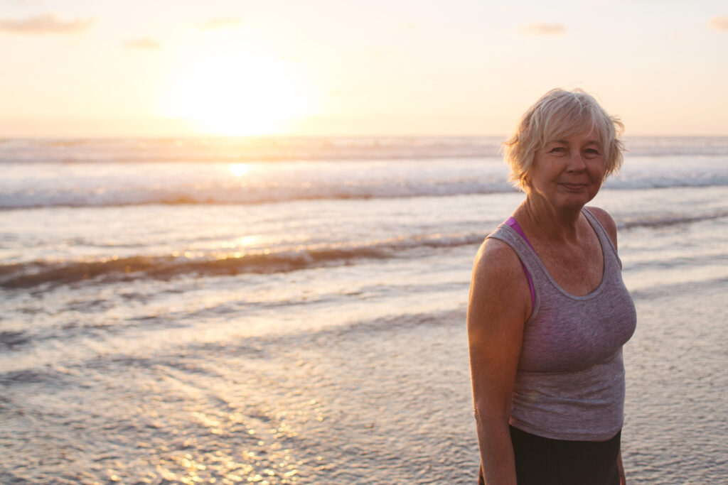 A smiling older woman in a tank top stands in front of an ocean sunset.