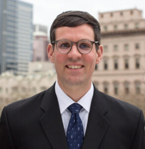 A smiling man in glasses and a suit stands in front of city buildings.