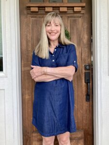 A smiling blonde woman in a blue dress stands in front of a wooden door.