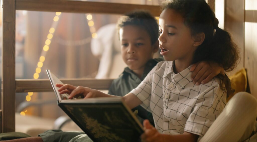 A young brother and sister sit by a window reading a book together.
