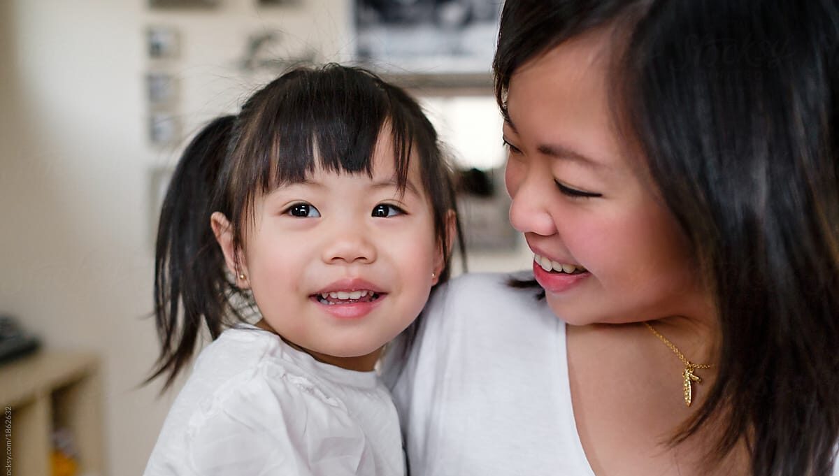 A smiling Asian woman looks at her young daughter, who she holds in her arms.