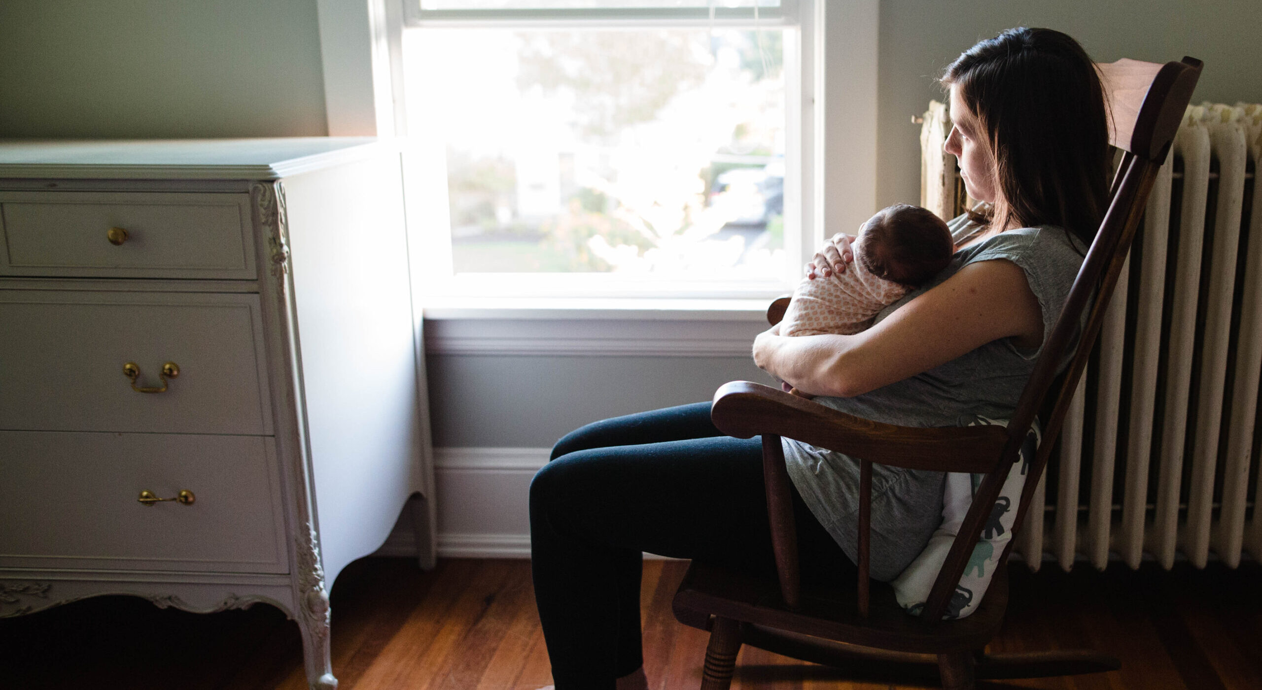 A young mother sits in a rocking chair looking out the window holding her infant in her arms. A heater is behind her.
