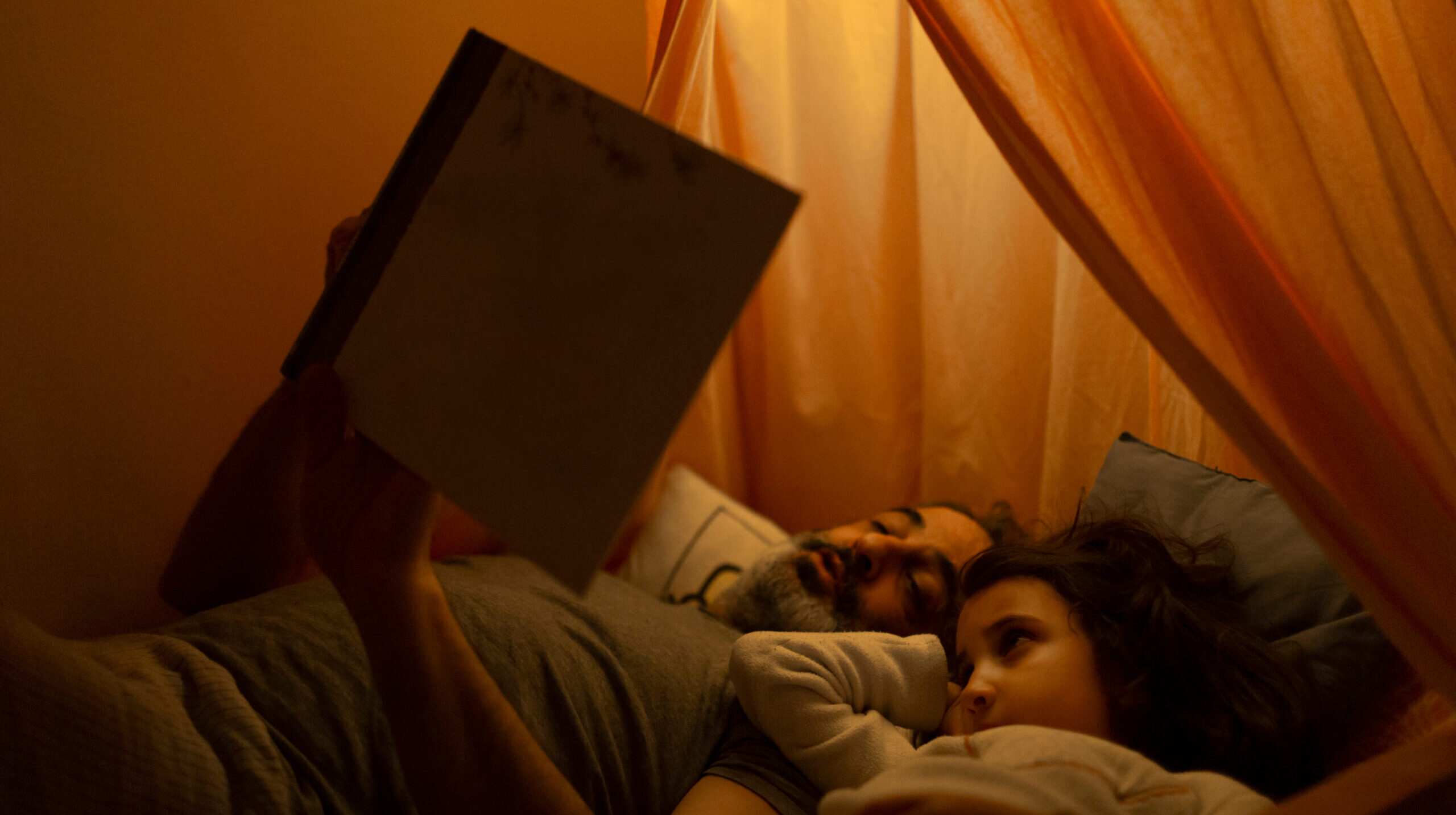 A father reads a book to his daugher in bed. They're under a pink tent in a kids' bedroom under fairy lights in their pajamas.