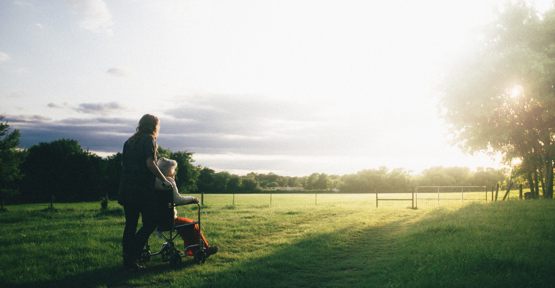 A caretaker pushes an elderly woman in a wheelchair across a sunny field